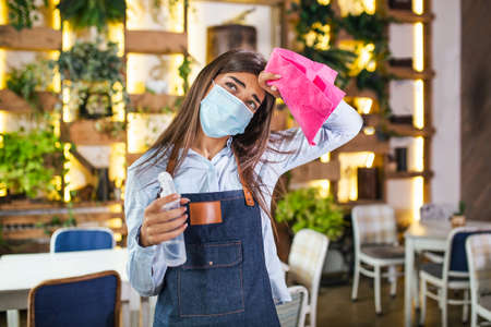 Portrait of attractive female waitress wearing face mask holding a bottle with sanitizer and a rag in a restaurant. New normal with coronavirus outbreak hygiene restaurant concept.の写真素材