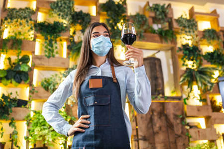 Young beautiful waitress wearing apron holding a glass of red wine in one hand serving a customer in a rustic restaurant. Sommelier recommended wine in restaurant.の写真素材