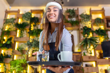 Female waitress wearing face shield, visor serves the coffee in restaurant during coronavirus pandemic representing new normal conceptの写真素材