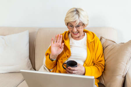 Enthusiastic progressive senior woman waving her hand and feeling happy while using a video call on laptop and talking with grandchildren. Elderly woman sitting on the sofa,holding cup of coffeeの写真素材