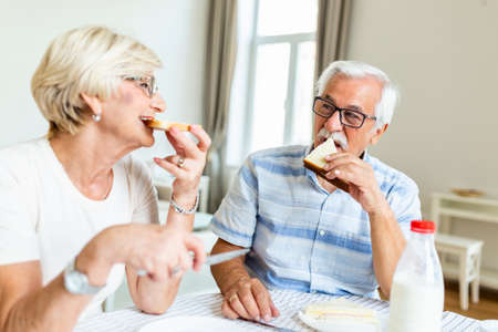 Senior couple eating breakfast at home. Holding piece of bread. Elderly couple enjoy in their time together. Feeling happy. Senior man eating sandwichの写真素材