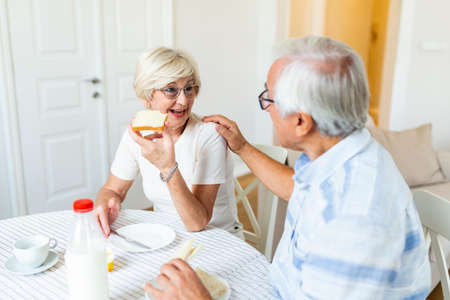 Cheerful senior couple having breakfast at home. Elderly man hugging woman. Mature woman holding...