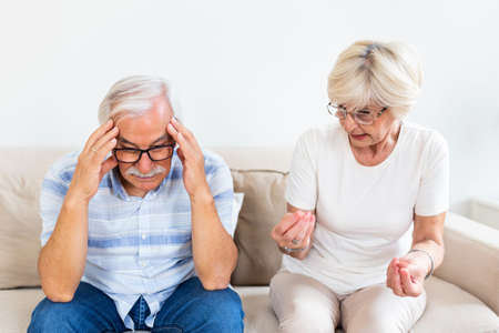 Elderly couple arguing. Senior man having headache, holding his head between hands. Mature woman frustrated with her husband. Marrige problemsの写真素材