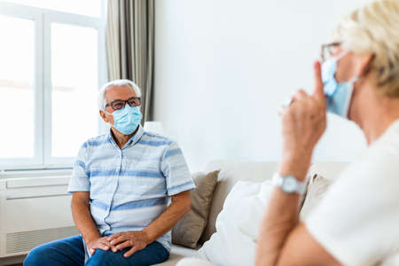 Senior friends having conversation on the sofa at home during Covid 19 pandemic. Elderly wearing medical face mask, preventing covid 19 coronavirus pandemic infection spread. Social distancingの写真素材