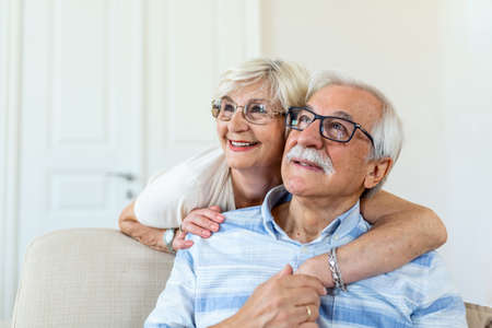 Portrait of overjoyed elderly husband and wife sit on couch hugging looking in distance. Happy mature spouses couple embrace cuddle relax together on sofa, enjoy time togetherの写真素材