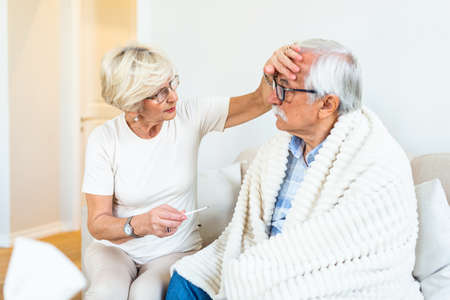 Woman checking fever temperature of senior man. Old husband resting at home feeling sick while his wife checking fever by touching foreheadの写真素材