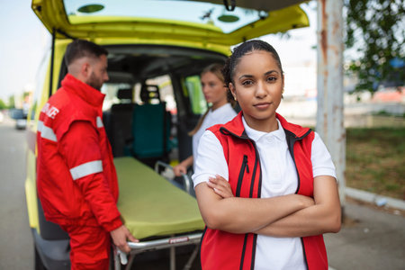 Young female african american paramedic smiling, standing rear of the ambulance, arms crossed. Three...