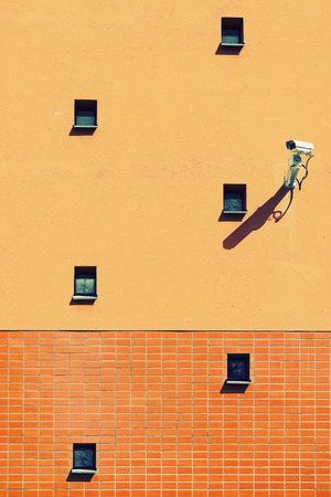 Surveillance camera on orange wall with small windows, filter applied for graphic effectの写真素材
