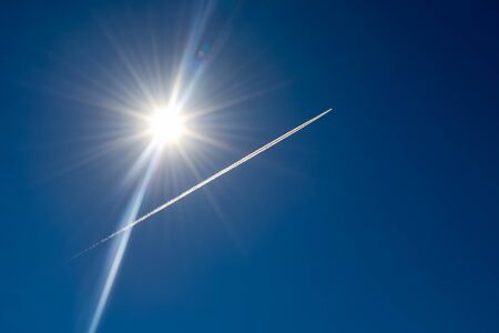 Condensation trail from a aircraft also called chemtrails against the bright sunの写真素材