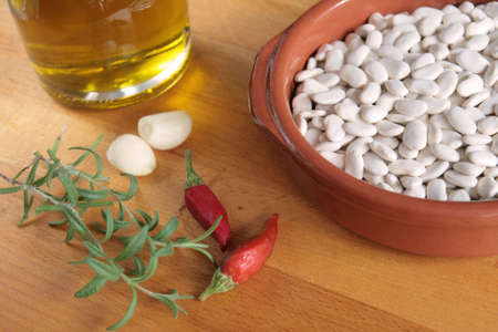 A bowl of raw haricot beans, with olive oil, garlic, chilli peppers and fresh rosemary on a wooden boardの写真素材