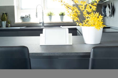 View of the modern kitchen. Mockup of a tablet with white blank screen over the kitchen table. Copy space. In the foreground branches of forsythia in a white vase, in the background window illuminated by natural sunlight. Italian interior designの写真素材