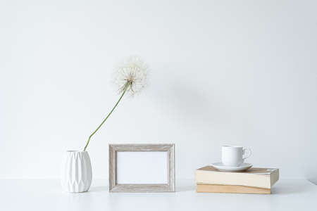 Empty wooden picture frame mockup. White vase with shower head, books and cup of coffee. Elegant lifestyle. White table and wall background. Modern, minimal and bright still lifeの写真素材