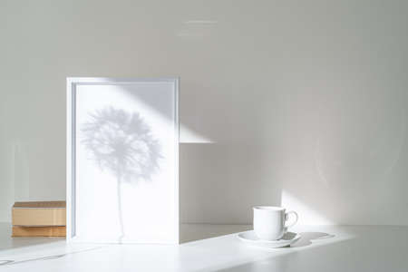 Empty white picture frame mockup in sunlight. Long shadow effect of shower head in the frame. Books and cup of coffee. Elegant lifestyle. White table and wall background illuminated by sun beam. Modern, minimal and bright still lifeの写真素材
