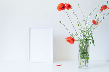 Empty white picture frame mockup. Glass vase with beautiful wild poppy flowers on white table. Lively and cheerful summer still life scene. Happy day conceptの写真素材