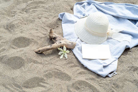 Summer still life. White straw hat on the sand of the mediterranean beach. Summer party invitation card mockup with white flower, branch and blue towel. Concept of vacation and relaxation. Business card and advertising for travel agencyの写真素材