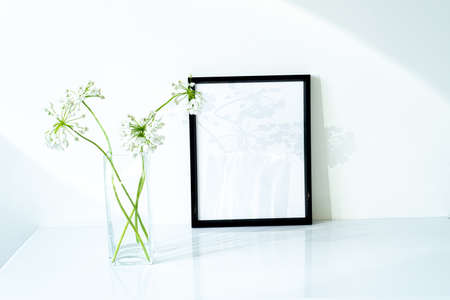Blank black frame mockup. Small fresh flowers of white garlic (allium neapolitanum) in glass vase in sunlight with long soft shadows. Glossy white table and white wall, minimal and elegant spaceの写真素材