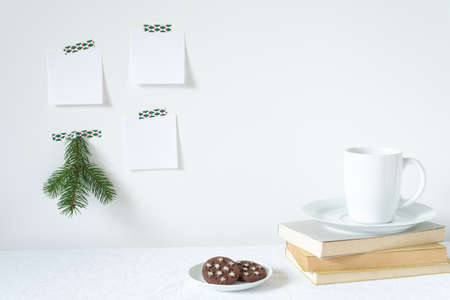 Festive breakfast still life.Cup of coffee with cookies, books on desk.Fir branch, empty notepads mockup hanging on the white wall.Christmas decor.Working space, home office.Happy new year resolutions, listの写真素材