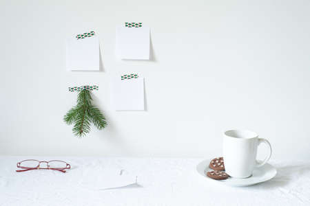 Festive breakfast still life.Cup of coffee with cookies, glasses on desk.Fir branch, empty notepads mockup hanging on the white wall.Christmas decor.Working space, home office.Happy new year resolutions, listの写真素材