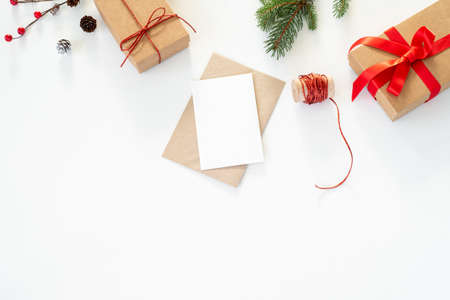 Christmas composition. Blank greeting card, envelope mockup.Decoration with christmas gifts with red bow, red berries, ribbon, fir tree branches and pine cones on white table background.Flat lay, top viewの写真素材