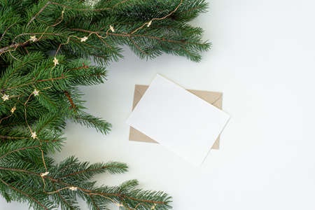 Christmas composition. Blank greeting card, envelope mockup.Fir tree branches and Christmas decorative lights on white table background.Flat lay, top viewの写真素材