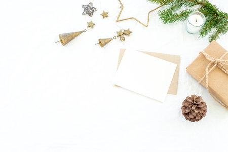 Christmas decorations composition. Christmas greeting card mock-up scene. Blank greeting card and envelope, gift christmas box with ribbon, fir branch, pine cone, metal decorations and burning candle. White linen background.Copy space, top view, flat layの写真素材