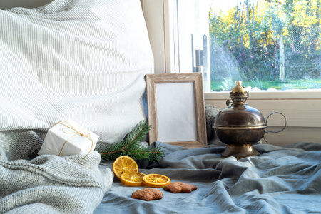 Cozy Christmas scene near the window. Winter interior. Blank wooden picture frame mockup. Wholemeal cookies, pillow, wool sweater, vintage wick oil lamp, gift box and fir tree branch.の写真素材