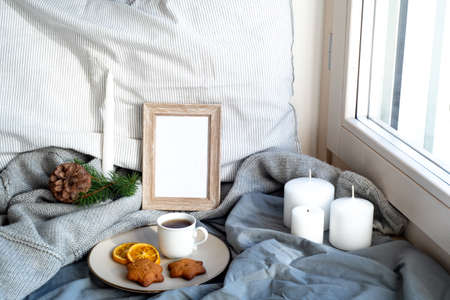 Cozy Christmas breakfast still life scene near the window. Winter interior. Blank wooden picture frame mockup.Cup of coffee with wholemeal cookies, pillow, wool sweater, candle, fir tree branch.の写真素材
