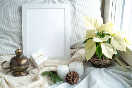 Cozy winter home interior scene.Christmas composition near the window.Blank white pictur frame mockup, white poinsettia, oil lamp, candles, gift box.Model for Christmas menu.の写真素材