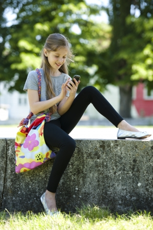 Little girl sending text messaging with mobile phone while sitting outdoorsの写真素材