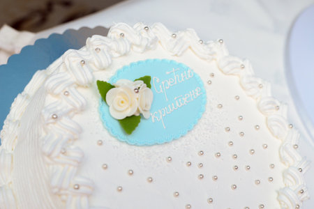 Part of a white round cake with a blue decoration and a white flower next to which is written in Cyrillic Happy Baptismの写真素材