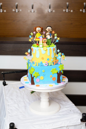 A colorful two-tiered birthday cake with the name Lazar written in Cyrillic, figures in the shape of mom, dad, baby and dog and a candle in the shape of the number oneの写真素材