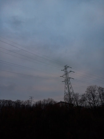 Silhouette of trees without leaves as it is winter time behind which the roof of some house is being monitored and above all that a power line with cables in cloudy weatherの写真素材