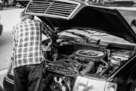 You see a black and white picture of an old car on a street with the bonnet open and the engine interior being repaired by a humanの写真素材