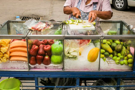 on the picture you can see a woman with her fruit cart made of glass and metal while she is cutting fruit for a customerの写真素材