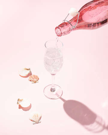 Creative celebration concept made of pink bottle pouring champagne in glass on light pink pastel table background with seashells. Artistic aesthetic.の写真素材