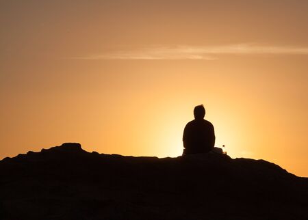 Silhouette of lone man sitting on a rock near the sea and watching the sunsetの写真素材