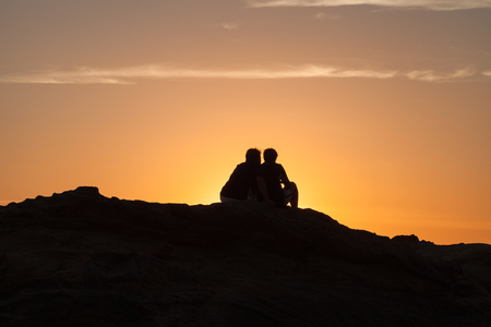 Silhouette of Couple sitting on a rock near the sea and watching the sunsetの写真素材