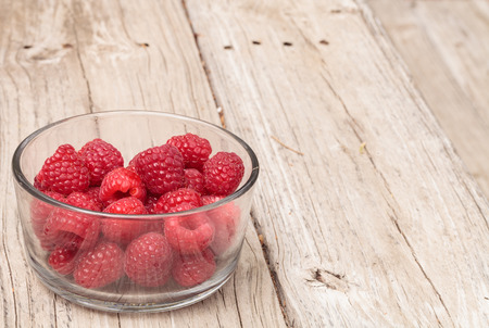 Clear glass bowl of ripe raspberries on a rustic farm picnic table in summer.