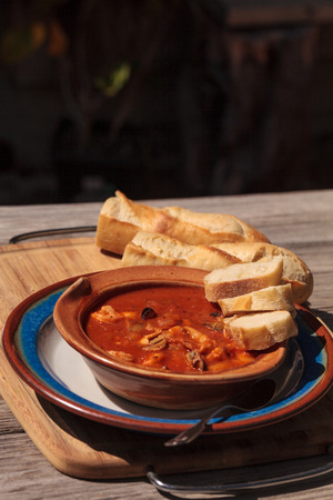Seafood cioppino with French bread and shrimp, fish, clams, lobster, crab and scallops in tomato sauce in a pottery bowl on a dish and a cutting board.の写真素材