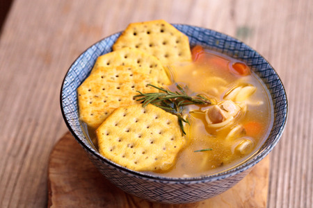 Chicken noodle soup in a blue and white bowl with crackers, all sitting on a wood cutting board.の写真素材