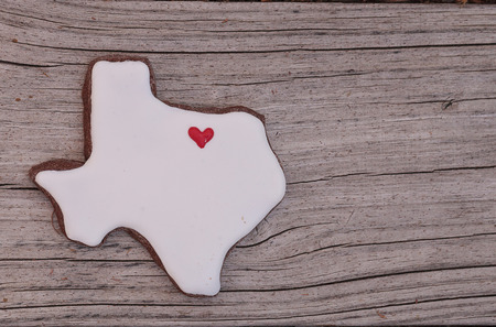 Texas shaped sugar cookie with white icing and a red heart on a rustic wood table.の写真素材