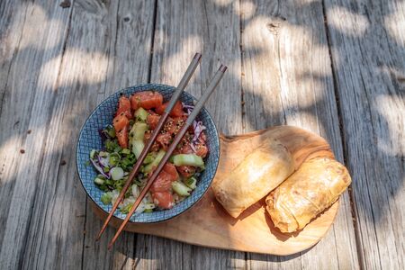 Raw salmon poke bowl with rice, cabbage, cucumber, sesame seeds and spring rolls with chopsticks.