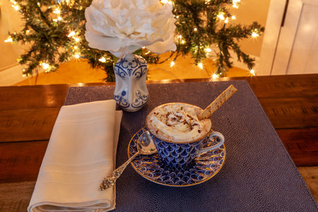 Cup of Hot chocolate in a fancy bone China cup with gold and a silver spoon in front of a Christmas tree.の写真素材
