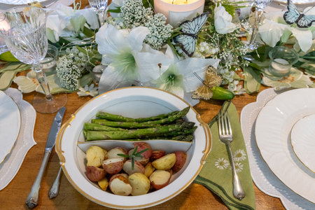 Asparagus and small roasted potatoes in a porcelain dish rimmed with gold on an Easter decorated table setting with lilies, butterflies, crystal and candles.の写真素材