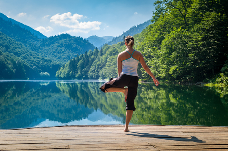 Young woman is doing yoga exercises at wooden pier. Beautiful nature landscape with forests on the mountains and mirror waters of the lake. Biogradska gora national park, Montenegro.の写真素材