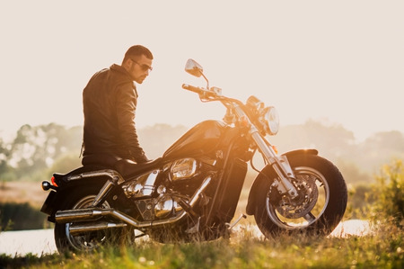 young brutal man in a black jacket and glasses standing near a motorcycleの写真素材