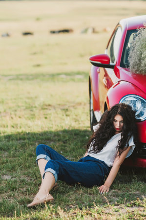 Beautiful young woman with long curly hair sitting near the red carの写真素材