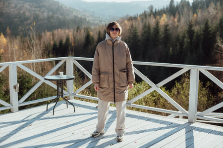 A young man in a brown jacket and sunglasses stands on a wooden terrace in the mountains.の写真素材