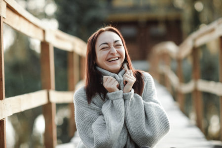 Happy young woman in sweater and scarf sitting on wooden bridge in winter park