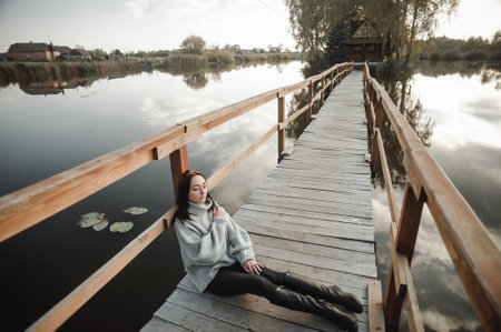 Young beautiful brunette girl in a gray sweater sits on a wooden bridge over the river.の写真素材